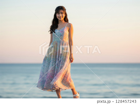 夏の夕暮れファッション：夕方の海辺で、ふんわりとした花柄キャミソールワンピースを着た女性モデル 128457243