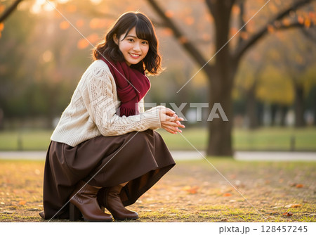 温もり感じる秋ファッション！夕日に照らされた公園で、深紅のマフラーを巻いて微笑む女性モデル 128457245