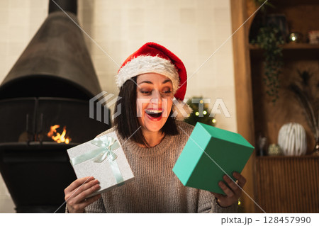 Merry Christmas. Young woman in Santa hat unpacking open up gift box near Christmas tree. Girl in living room with Christmas tree, fireplace opening gift box with surprise face. Christmas eve at home 128457990