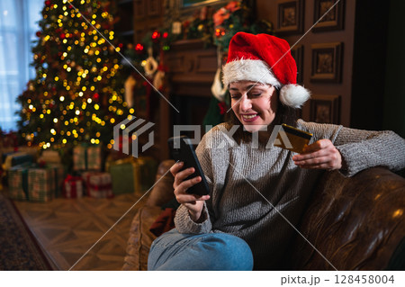 Merry Christmas. Young woman shopping online holding smartphone paying with gold credit card for Christmas gifts. Girl buying on Internet enter credit card details enjoying winter holidays at home Merry Christmas. Young woman shopping online holding smartphone paying with gold credit card for Christmas gifts. Girl buying on Internet enter credit card details enjoying winter holidays at home 128458004
