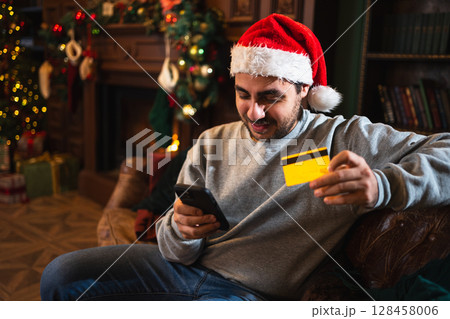 Merry Christmas. Man shopping online holding smartphone paying with gold credit card for Christmas gifts. Young adult man buying on Internet enter credit card details enjoying winter holidays at home 128458006