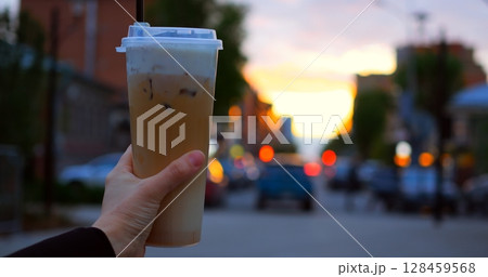 Female hand holding frothy cold coffee beverage, standing amid urban street during golden hour, blurred cityscape creating soft luminous backdrop 128459568