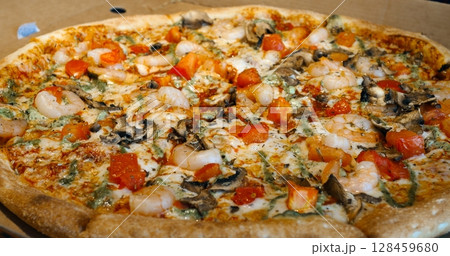 Round pizza with shrimps, tomatoes, mushrooms, and pesto rotates slightly on a cardboard box, showcasing its delicious toppings and crispy crust Round pizza with shrimps, tomatoes, mushrooms, and pesto rotates slightly on a cardboard box, showcasing its delicious toppings and crispy crust 128459680