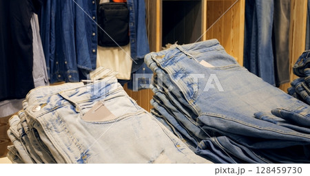 Multiple stacks of folded blue jeans sit on a wooden display table in a clothing store, surrounded by other apparel items, creating visually appealing display for shoppers. Concept fashion clothes Multiple stacks of folded blue jeans sit on a wooden display table in a clothing store, surrounded by other apparel items, creating visually appealing display for shoppers. Concept fashion clothes 128459730