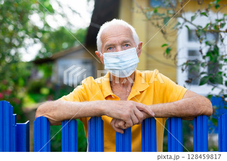 Portrait of gray-haired owner in protective mask at fence of his country house in village Portrait of gray-haired owner in protective mask at fence of his country house in village 128459817