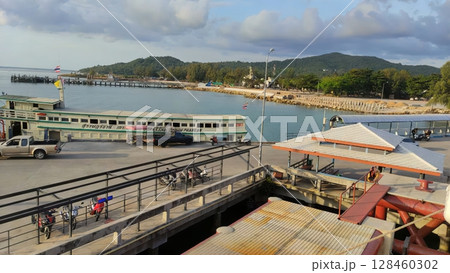 Green and white ferry navigating toward pier on koh chang island, thailand, with parked motorbikes waiting under overcast sky, preparing for maritime passenger transportation 128460302