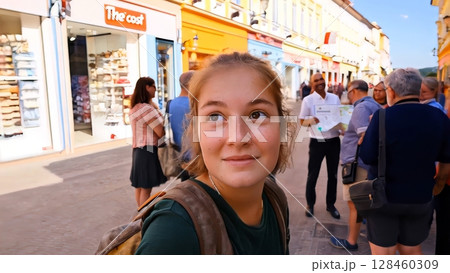 Tourist girl listening with interest to the guide's explanations, during a guided tour in a picturesque european city, with colorful buildings and shops 128460309