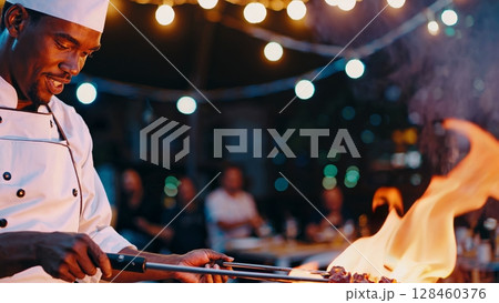 African american chef grilling meat skewers over a barbecue fire at an outdoor evening event, illuminated by string lights, demonstrating culinary expertise and passion for food 128460376