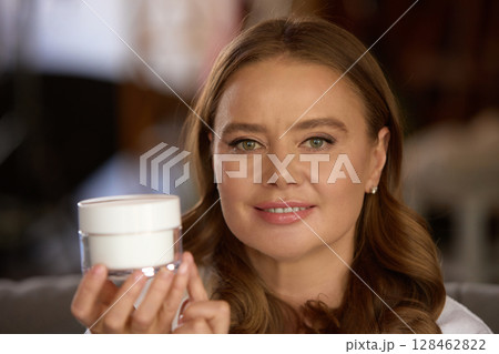 Smiling mature woman in white robe holding face cream jar in warm natural light Smiling mature woman in white robe holding face cream jar in warm natural light 128462822