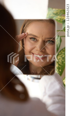 Smiling mature woman checks face in mirror under daylight at home Smiling mature woman checks face in mirror under daylight at home 128462823
