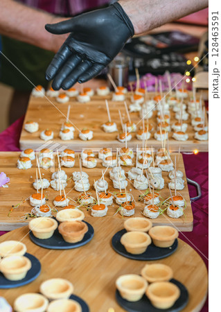 Mini canapes on wooden boards with toothpicks and chefs gloved hand presenting appetizers at food event. Concept of catering, finger food and culinary presentation 128463591