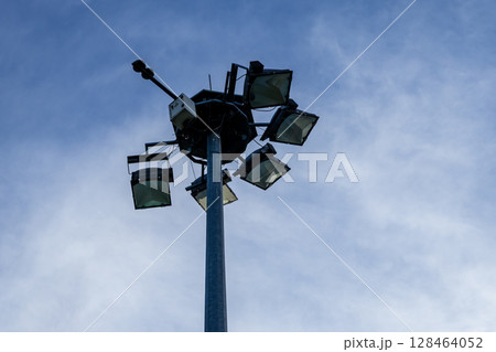 A powerful high mast lighting system with multiple bright floodlights on a tall pole against a blue sky, providing extensive illumination for large outdoor public areas A powerful high mast lighting system with multiple bright floodlights on a tall pole against a blue sky, providing extensive illumination for large outdoor public areas 128464052