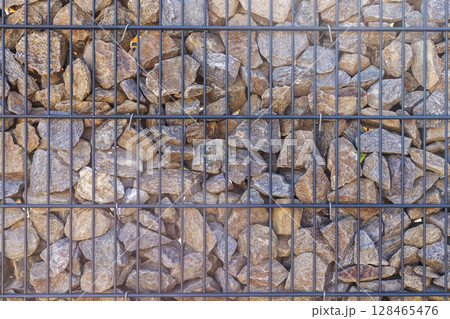 A detailed view of a wall made of irregularly shaped rocks secured within a black metal grid. The stones vary in size and color, creating an organic pattern 128465476
