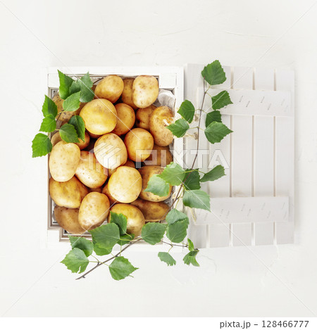 Raw potatoes. Natural, farm potatoes in a white wooden box with green leaves on white background. Selling seasonal vegetables at local market. Support local farmers and harvesting concept. Flat lay Raw potatoes. Natural, farm potatoes in a white wooden box with green leaves on white background. Selling seasonal vegetables at local market. Support local farmers and harvesting concept. Flat lay 128466777