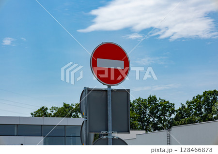 A bright red no entry sign is visible against a clear blue sky, surrounded by greenery and a modern building in the background 128466878