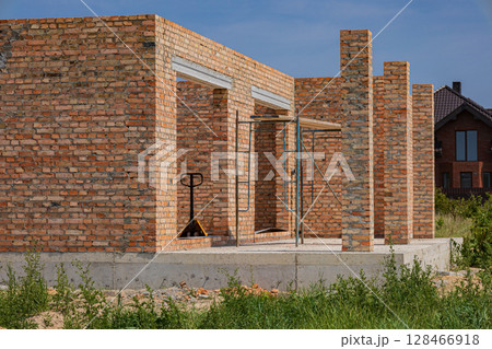 Brick walls are being constructed at a building site, with a pallet jack visible, surrounded by greenery under a clear blue sky 128466918