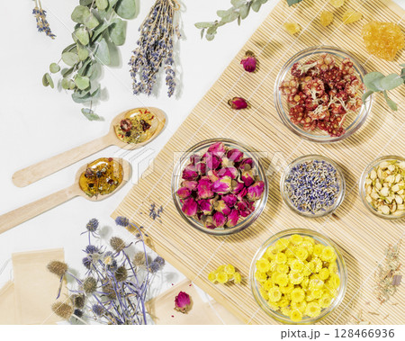 Home herbal apothecary concept. Sustainable flat lay with dry flowers and herbs, tea bags and wooden spoons on wooden napkin on white background. Healing tea from wild plants and flowers. 128466936