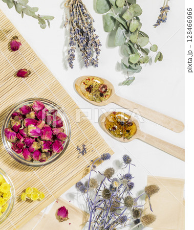 Home herbal apothecary concept. Dry flowers and herbs, tea bags and wooden spoons on wooden napkin on white background. Healing tea from wild plants and flowers. Flat lay, vertical format 128466966