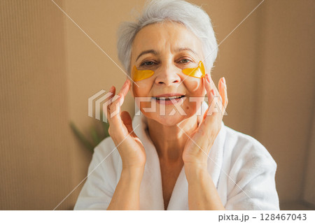 Eye patch beauty aging treatment. Mature adult old woman with under eye collagen pads. Senior woman with patches under eyes gets facial treatment. Senior lady in robe relaxing in bathroom at home 128467043