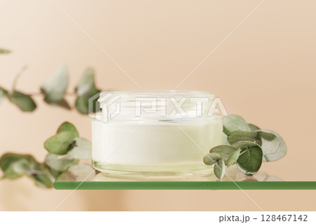Cosmetic glass jar with natural white cream on glass shelf on pink background with eucalyptus sprigs. Organic beauty product and skin friendly cosmetics concept. Mockup, front view, close up 128467142