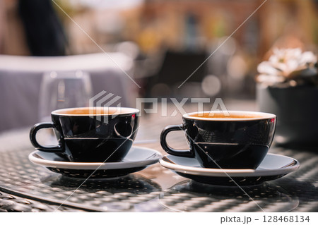 Two dark coffee cups sit on a table in a sunny outdoor cafe setting 128468134