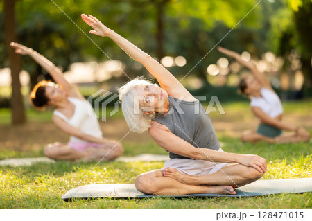 Sporty old lady sitting on sports mat and making yoga meditation in lotus pose and hands up with group of women together in green park at daytime Sporty old lady sitting on sports mat and making yoga meditation in lotus pose and hands up with group of women together in green park at daytime 128471015