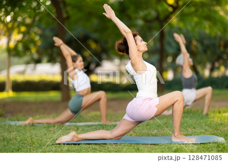 Sporty energetic female teacher doing exercises on pilates mat and demonstrating variation of warrior pose of yoga for group women at city park. Fitness concept Sporty energetic female teacher doing exercises on pilates mat and demonstrating variation of warrior pose of yoga for group women at city park. Fitness concept 128471085