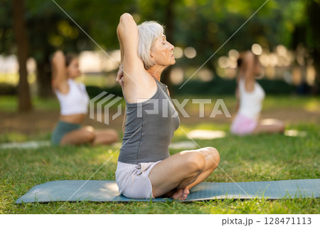 Sporty old lady sitting on sports mat and making yoga meditation in lotus pose and hands up with group of women together in green park at daytime 128471113