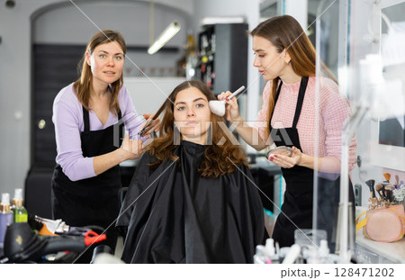 Professional female stylists making hairstyle and makeup to young woman 128471202