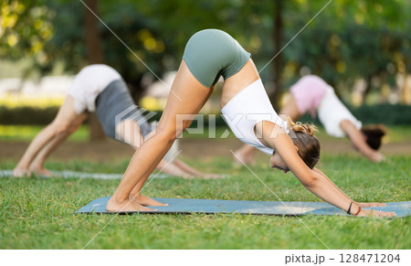 Girl doing Downward Facing Dog Pose during group yoga in park 128471204