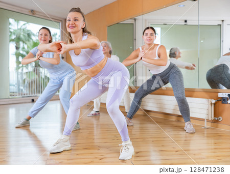 Four emotional fit young mature women in sportswear warming up with squats before aerobic dance training in gym Four emotional fit young mature women in sportswear warming up with squats before aerobic dance training in gym 128471238