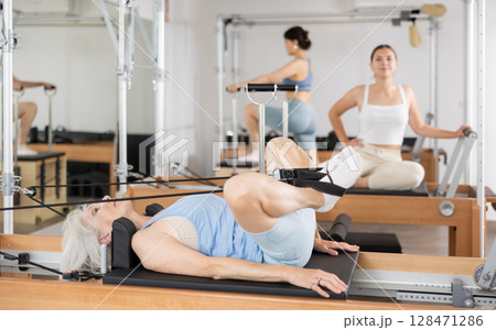 Elderly woman performing set of pilates exercises on reformer 128471286
