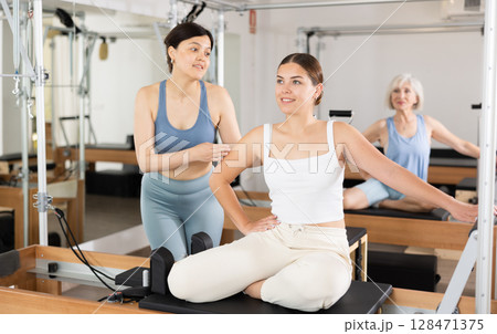 Asian female pilates trainer controlling movements of young woman on reformer 128471375
