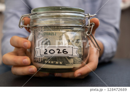 Unrecognizable woman holding Saving Money In Glass Jar filled with Dollars banknotes. 2026 year transcription in front of jar. Managing personal finances extra income for future insecurity Unrecognizable woman holding Saving Money In Glass Jar filled with Dollars banknotes. 2026 year transcription in front of jar. Managing personal finances extra income for future insecurity 128472689