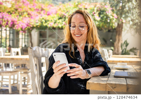 Smiling Woman Using Smartphone at Outdoor Cafe Table with Laptop and Coffee on a Sunny Day 128474634