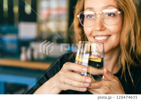 Happy Woman Holding Glass of Water with Lemon, Smiling in Cozy Cafe, Healthy Lifestyle, Relaxation Happy Woman Holding Glass of Water with Lemon, Smiling in Cozy Cafe, Healthy Lifestyle, Relaxation 128474635