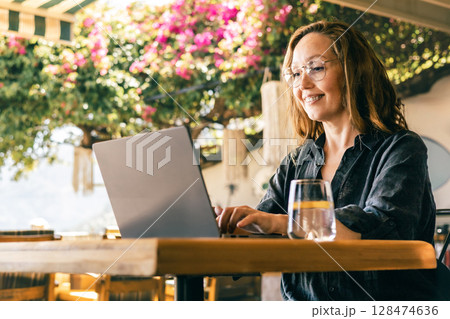 Happy Woman Working on Laptop Outdoors Under Blooming Tree in Sunny Cafe Environment Happy Woman Working on Laptop Outdoors Under Blooming Tree in Sunny Cafe Environment 128474636