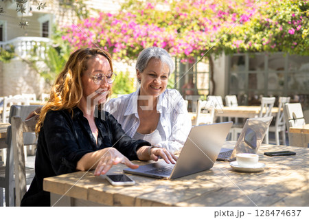 Smiling Women Working Together on Laptops Outdoors in Sunny Cafe, Remote Team Collaboration Smiling Women Working Together on Laptops Outdoors in Sunny Cafe, Remote Team Collaboration 128474637