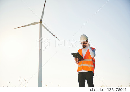 In sunglasses, with notepad. Service engineer is on the field with windmills In sunglasses, with notepad. Service engineer is on the field with windmills 128478041
