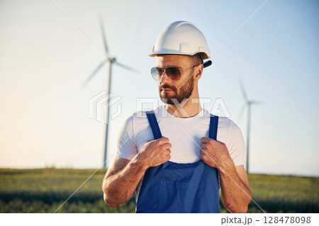 In blue colored uniform and in sunglasses. Service engineer is on the field with windmills 128478098