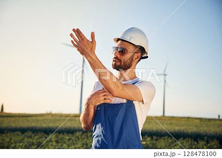 In blue colored uniform and in sunglasses. Service engineer is on the field with windmills 128478100
