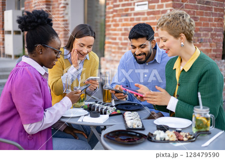A diverse group of friends are gathered at an outdoor cafe, enjoying food and using their smartphones. A diverse group of friends are gathered at an outdoor cafe, enjoying food and using their smartphones. 128479590