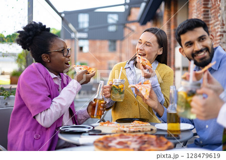 A group of friends enjoys pizza and drinks outdoors, laughing and sharing a meal together. A group of friends enjoys pizza and drinks outdoors, laughing and sharing a meal together. 128479619