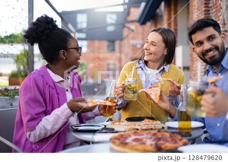 Group of friends enjoying pizza and drinks outdoors, laughing and having fun together on a sunny day. 128479620