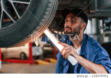 Under car, using lighting device. Auto mechanic working in garage. Repair service 128479701