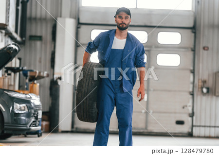 Front view, standing and holding tire. Auto mechanic working in garage. Repair service Front view, standing and holding tire. Auto mechanic working in garage. Repair service 128479780