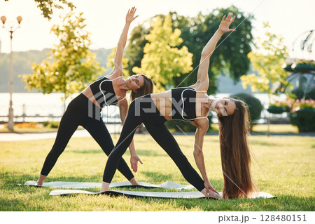 Doing yoga exercises. Two women in sport clothes are doing exercises outdoors on the field 128480151