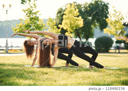 On the yoga mat. Two women in sport clothes are doing exercises outdoors on the field 128480156