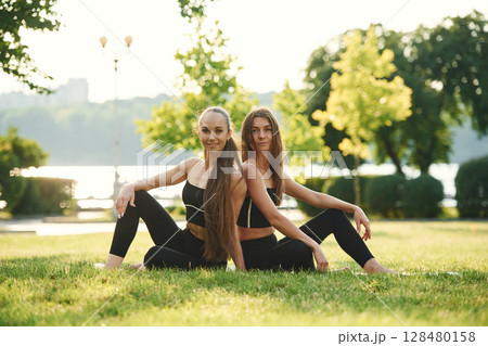 On the yoga mat. Two women in sport clothes are doing exercises outdoors on the field 128480158