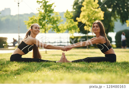 Holding each other by hands, touching feet. Two women in sport clothes are doing exercises outdoors on the field Holding each other by hands, touching feet. Two women in sport clothes are doing exercises outdoors on the field 128480163